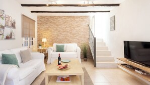 Un saló modern amb un sofà blanc, una taula de centre amb una ampolla i aperitius, una televisió sobre un suport de fusta, i una escala que condueix a un altre pis.