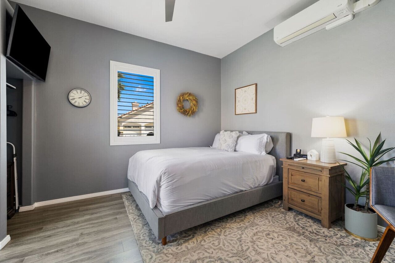 A bedroom with a bed, a nightstand, a wall clock, a window with blinds, a wreath, and a wall-mounted air conditioner.