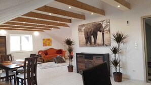 A spacious living room with a wooden ceiling, a dining area with a table and chairs, a sofa, and a fireplace. There are potted plants and a large elephant painting on the wall. - Excl. Apartment for the discerning, 75 sqm, attic, 2 pers. (Heeßen)