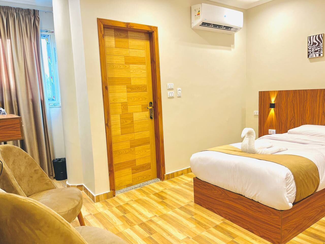 A hotel room with a wooden door, a bed with a white bedspread, a wooden headboard, and a small table with a lamp.