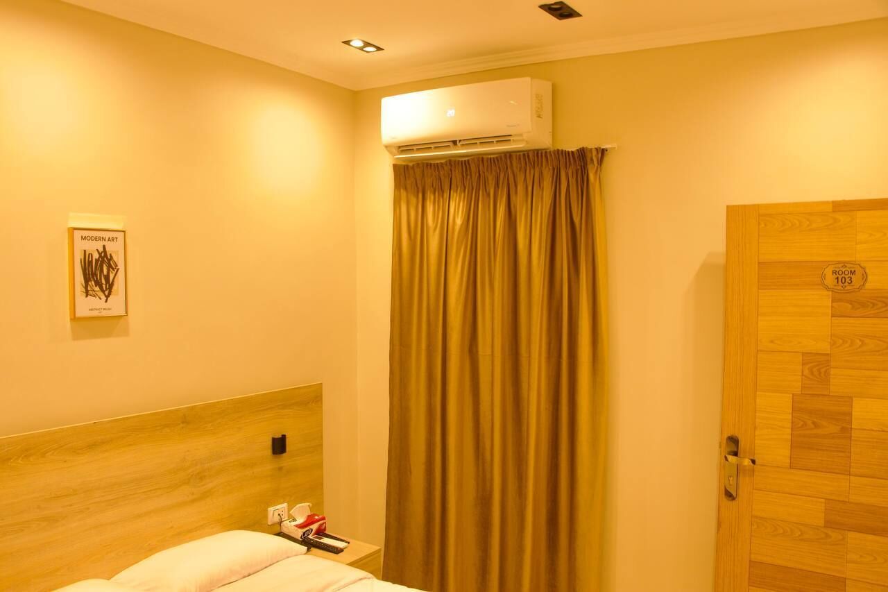 A hotel room with a wooden headboard, an air conditioning unit, a curtain, a door with a room number, and a framed artwork on the wall.