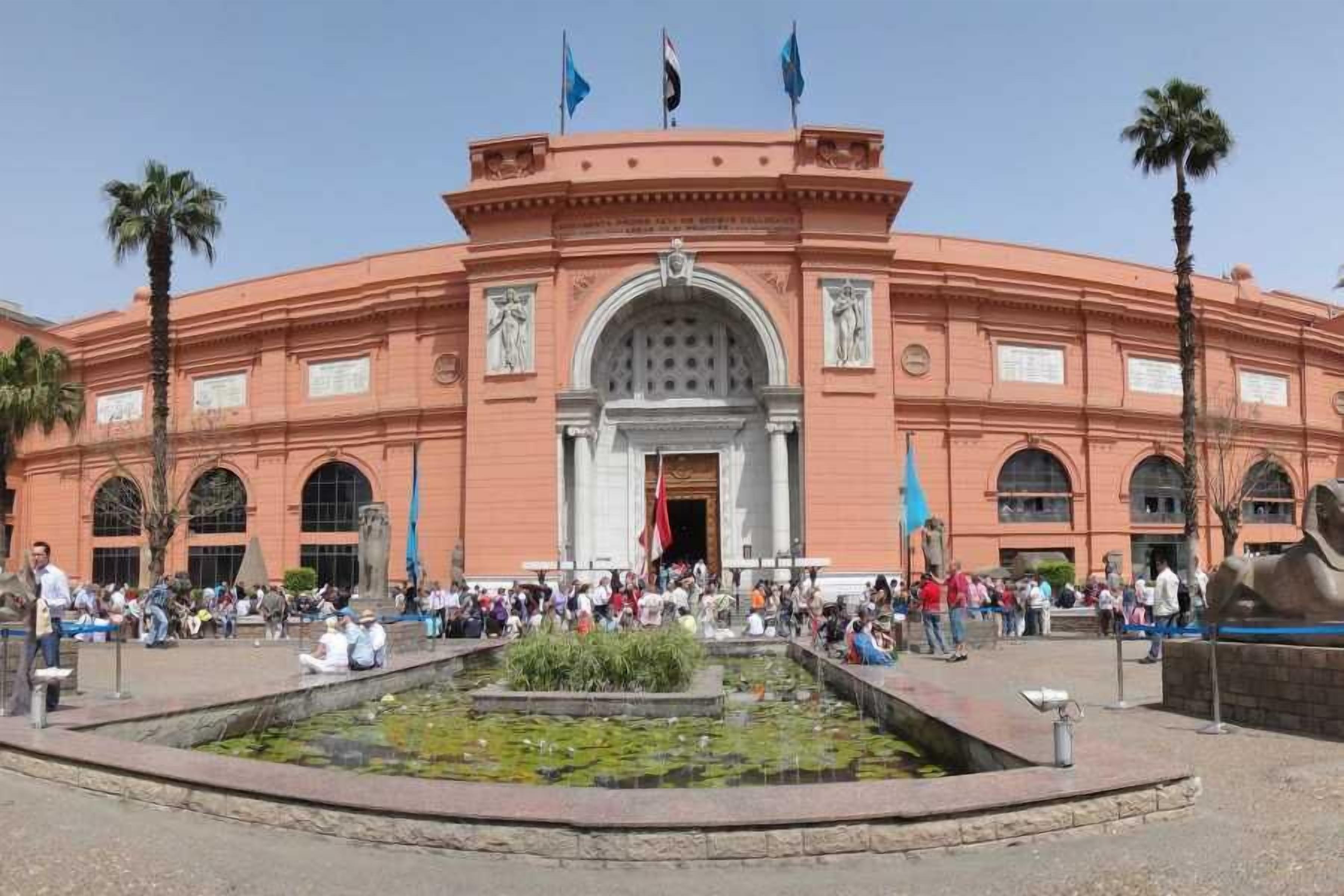 Un edificio grande e histórico con una entrada central arqueada, palmeras y una multitud de gente.