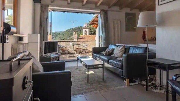 A living room with a sofa, coffee table, and a TV. There's a balcony with a view of a building and hills. - CASA  VISTA (Lugano)
