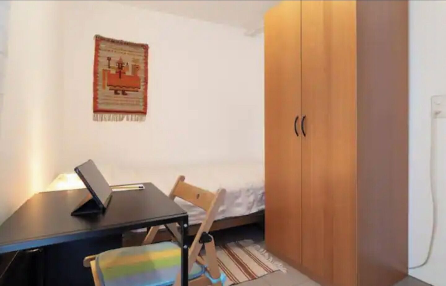 A small room with a bed, a wooden wardrobe, a desk with a laptop, and a chair.