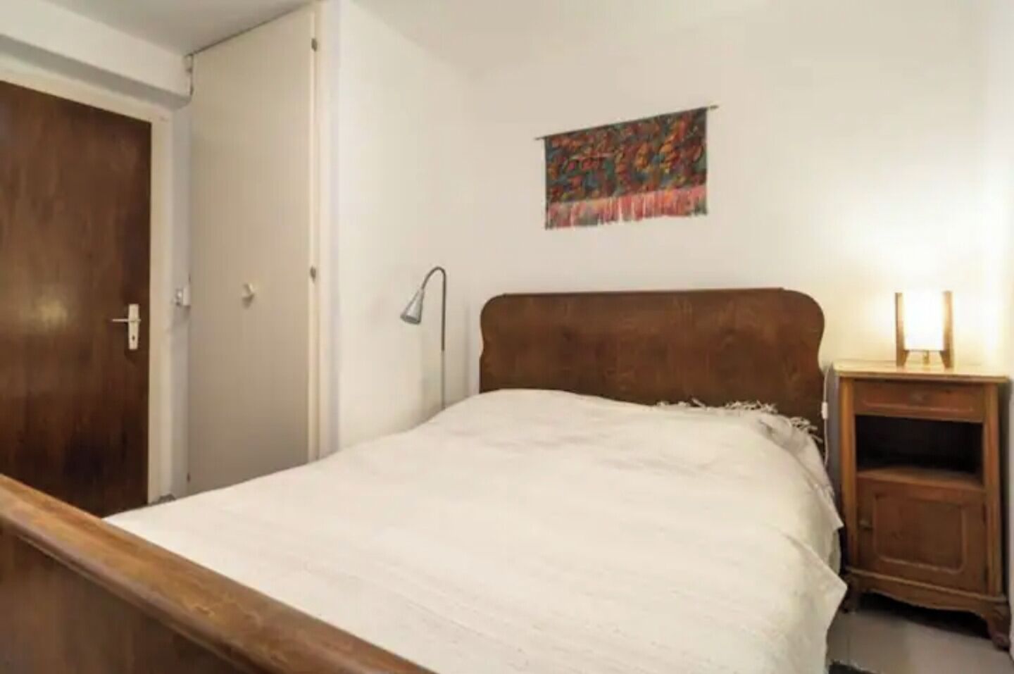 A bedroom with a wooden bed frame, a nightstand with a lamp, and a colorful painting on the wall.