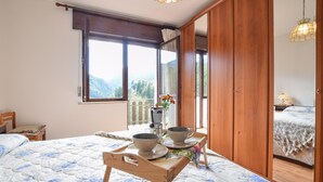 Een slaapkamer met een bed, een houten stoel, een tafel met kopjes en een bloevas, en een uitzicht op bergen door een raam.