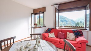 Een gezellige kamer met een rode bank, een eettafel met bloemrijk servies en uitzicht op bergen door grote ramen.