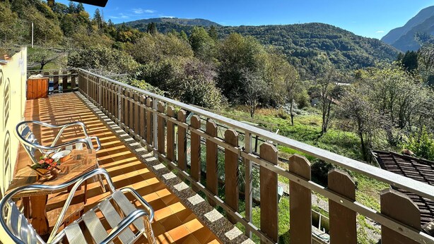 Een balkon met een houten vloer, metalen stoelen en uitzicht op een bergachtig landschap.