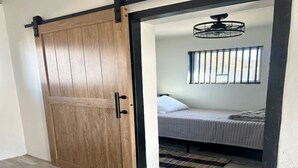 Una habitación con una puerta corredera de madera, una cama con una colcha a rayas, una ventana con barrotes y un ventilador de techo.