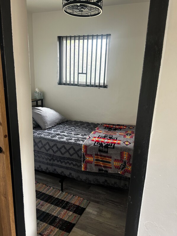 A small bedroom with a single bed, a window with vertical blinds, a bedside table with a tissue box, and a patterned rug on the floor.