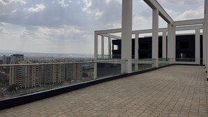 Un balcón amplio con suelo de baldosas y barandilla de cristal, con vistas a un paisaje urbano que incluye edificios y un cielo nublado.