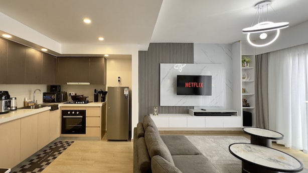 Una cocina moderna con horno empotrado, microondas y refrigerador. Hay un comedor con sofá y una mesa de centro. Un televisor de pantalla plana montado en la pared muestra
