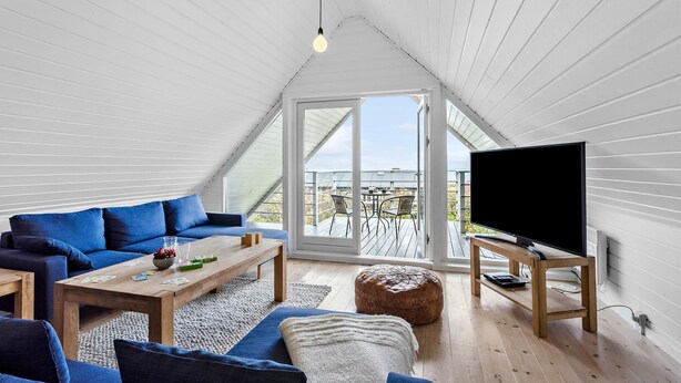 Ein modernes Loft-Zimmer mit schrägverdeckter Decke, einem großen Fernseher, einer Couch, einem Couchtisch und Blick ins Freie durch eine Glastür.