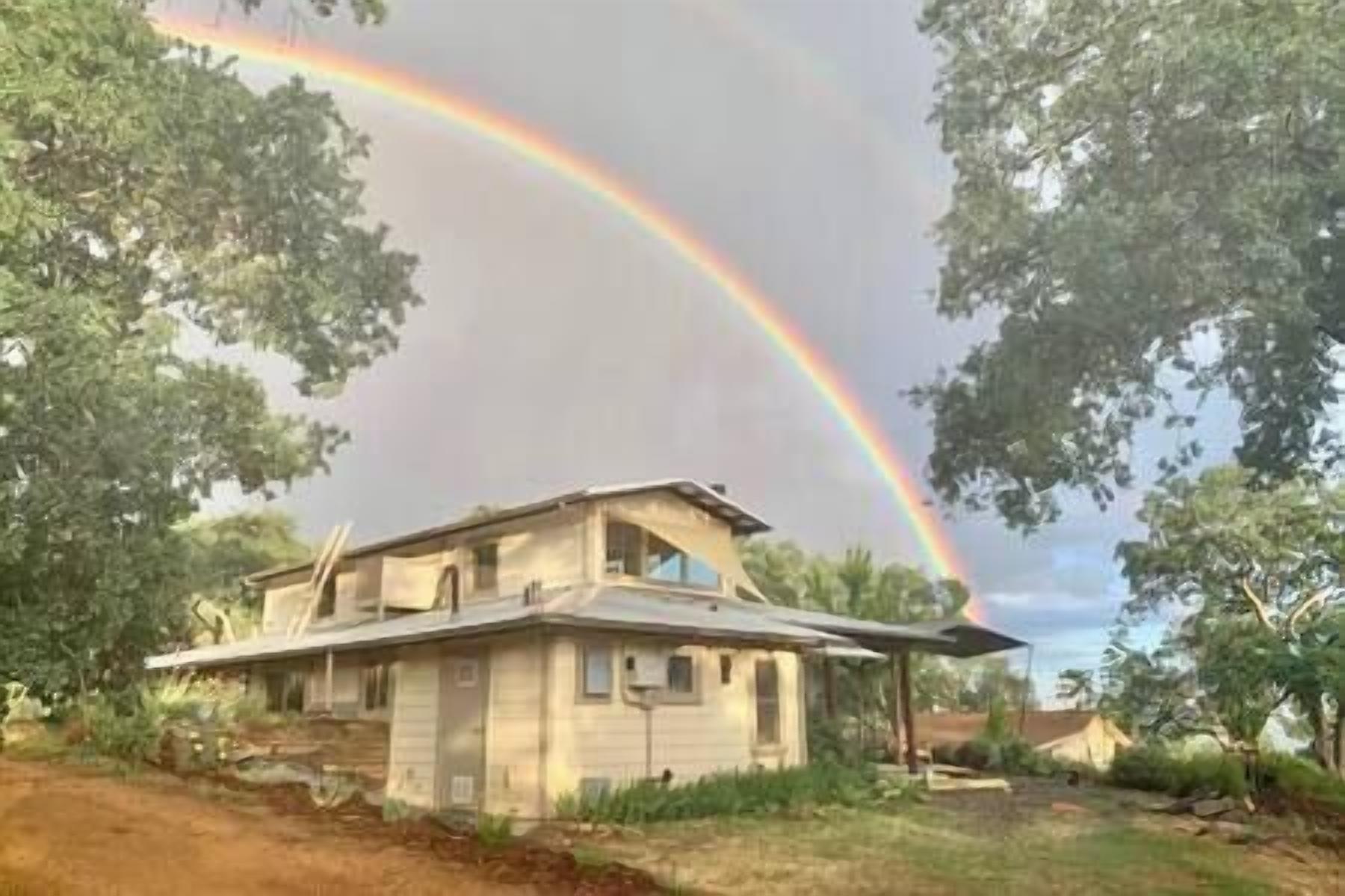 Une maison à deux étages, avec un toit en pente, entourée d’arbres et bordée d’un chemin de terre.