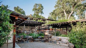 A stone patio with a pergola structure, surrounded by greenery and trees.