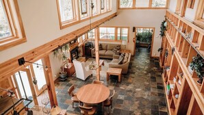 A spacious interior with a high ceiling, large windows, a central seating area with a sofa and chairs, a dining table, and wooden shelving with plants.