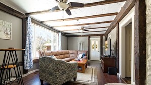 A spacious room with a high ceiling, exposed wooden beams, a ceiling fan, a large window, a sofa, a patterned armchair, a small table, and a door leading to another room.