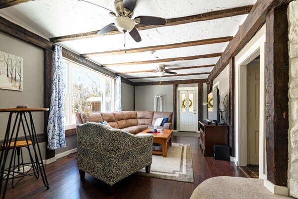 A spacious room with a high ceiling, exposed wooden beams, a ceiling fan, a large window, a sofa, a patterned armchair, a small table, and a door leading to another room. - Every Season Get Away (Alexandria)