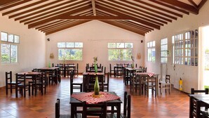 A spacious dining area with wooden beams on the ceiling, multiple tables with chairs, and large windows allowing natural light.