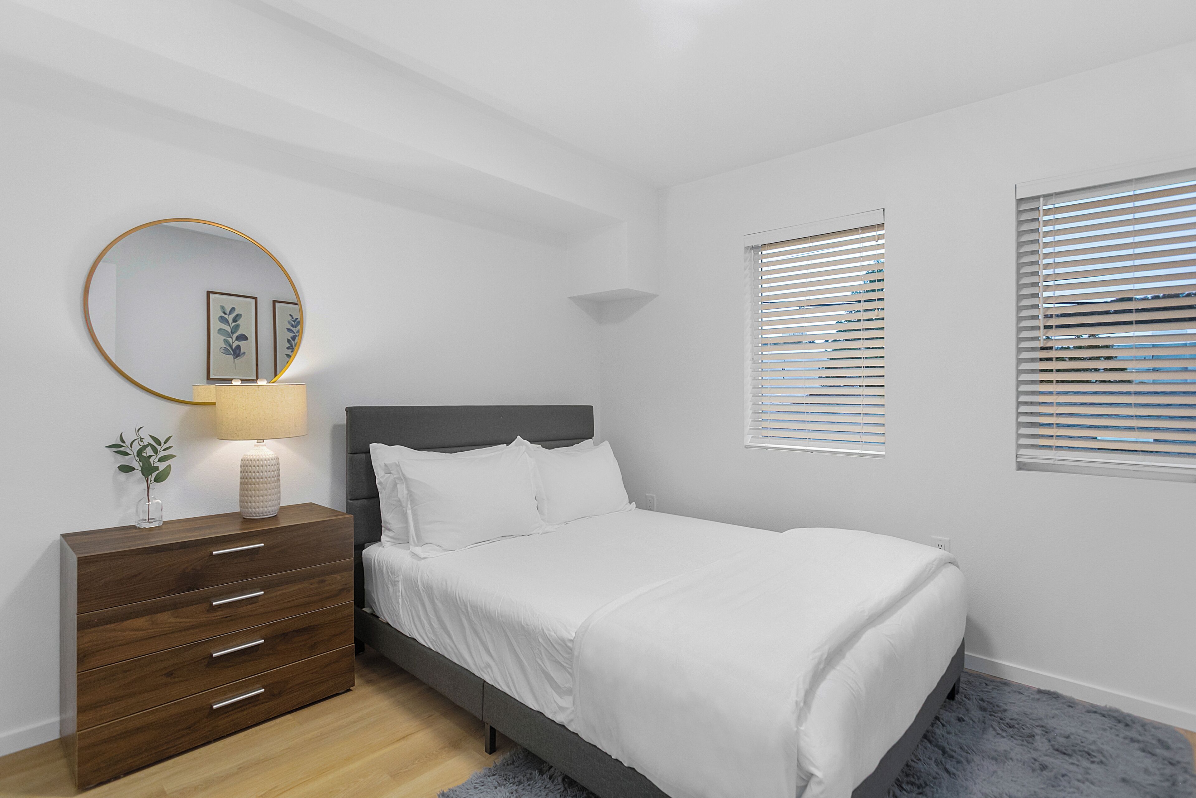 Un dormitorio con una cama, un aparador, una lámpara y dos ventanas con persianas.