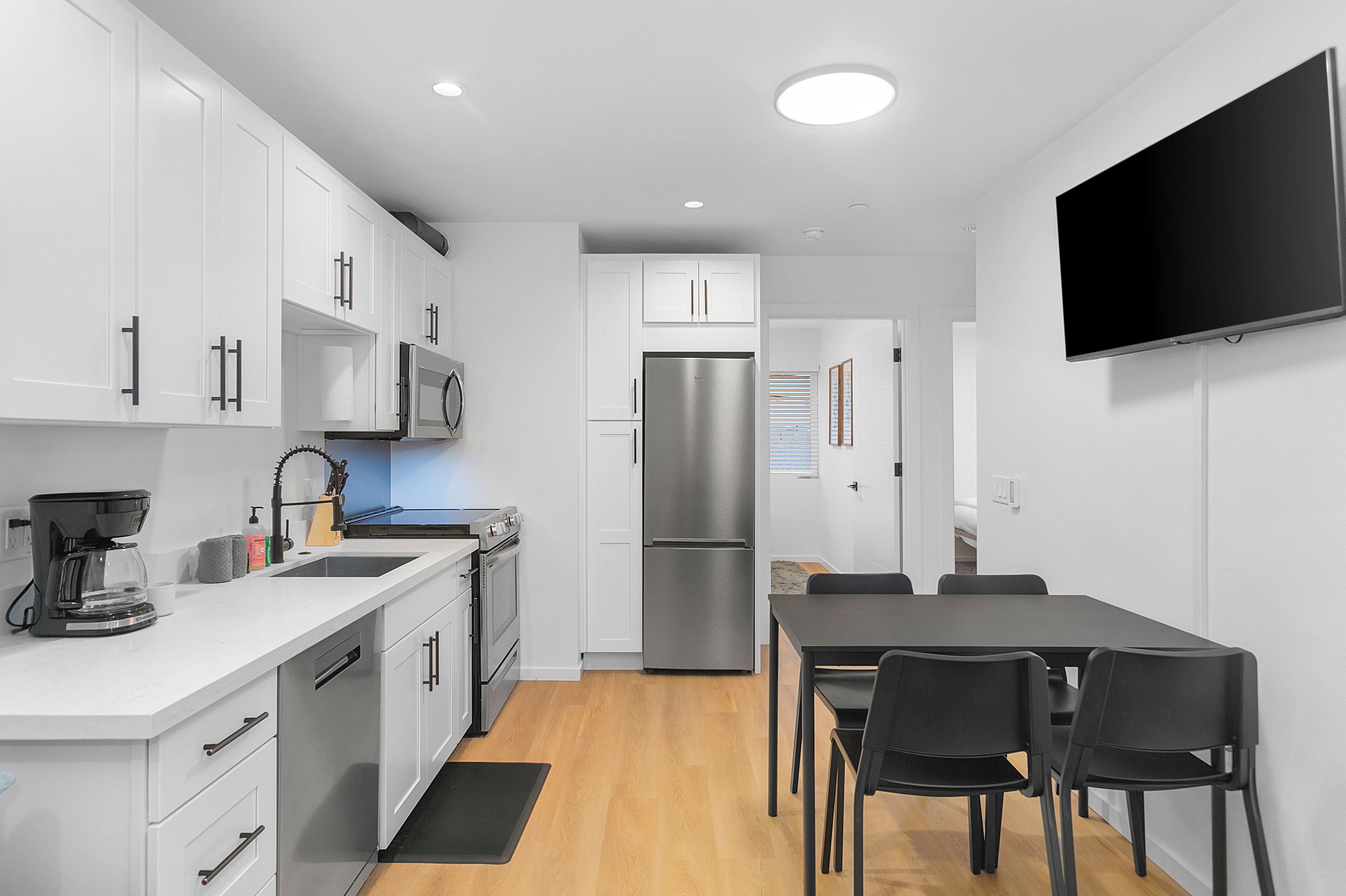 A modern kitchen with white cabinets, stainless steel appliances, a flat-screen TV, and a dining area with a table and chairs.