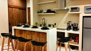A compact kitchen with a central island, bar stools, a built-in oven, a coffee maker, and a refrigerator. - Koom (Tulum)