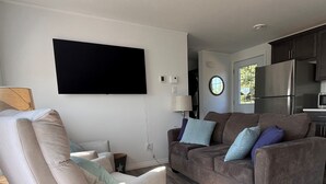 A modern living room with a sofa and armchair, a flat-screen TV, and a kitchen area in the background.