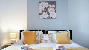 Una cama bien hecha con almohadas y una manta, una lámpara de noche y una obra de arte floral en la pared.