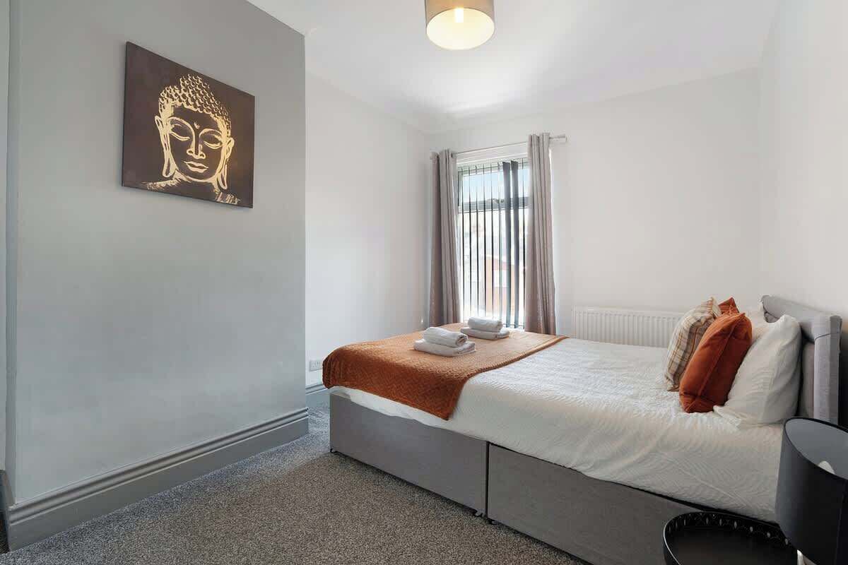 A bedroom with a bed, a headboard, a bedside table, a lamp, a window with curtains, and a wall-mounted artwork.
