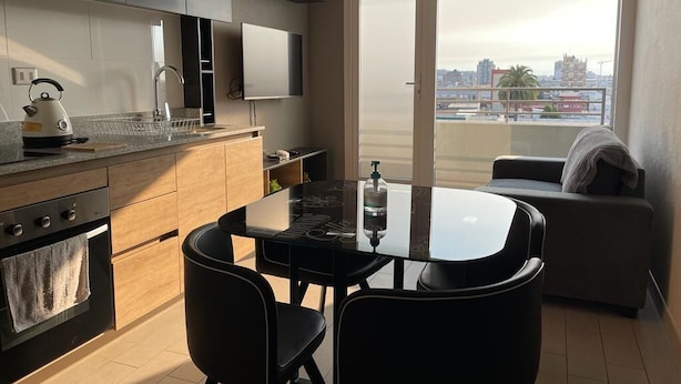 A modern kitchen with a dining area, a large window with a city view, and a sofa by the window. - Cozy fully equipped apartment in the center of Concepción. (Concepción)