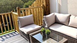 Un canapé en osier avec des coussins, une table en verre et des plantes en pot sur une terrasse en bois offrant une vue sur les arbres.