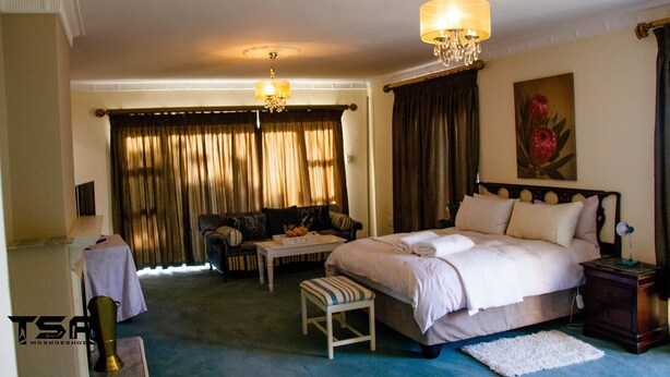 A hotel room with a large bed, a small table, a chair, and a nightstand. - The Advocate Suites Manor (Maseru)