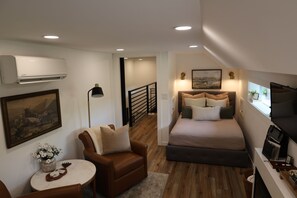 A bedroom with a bed, a chair, a table, an air conditioner, a painting, and a TV.