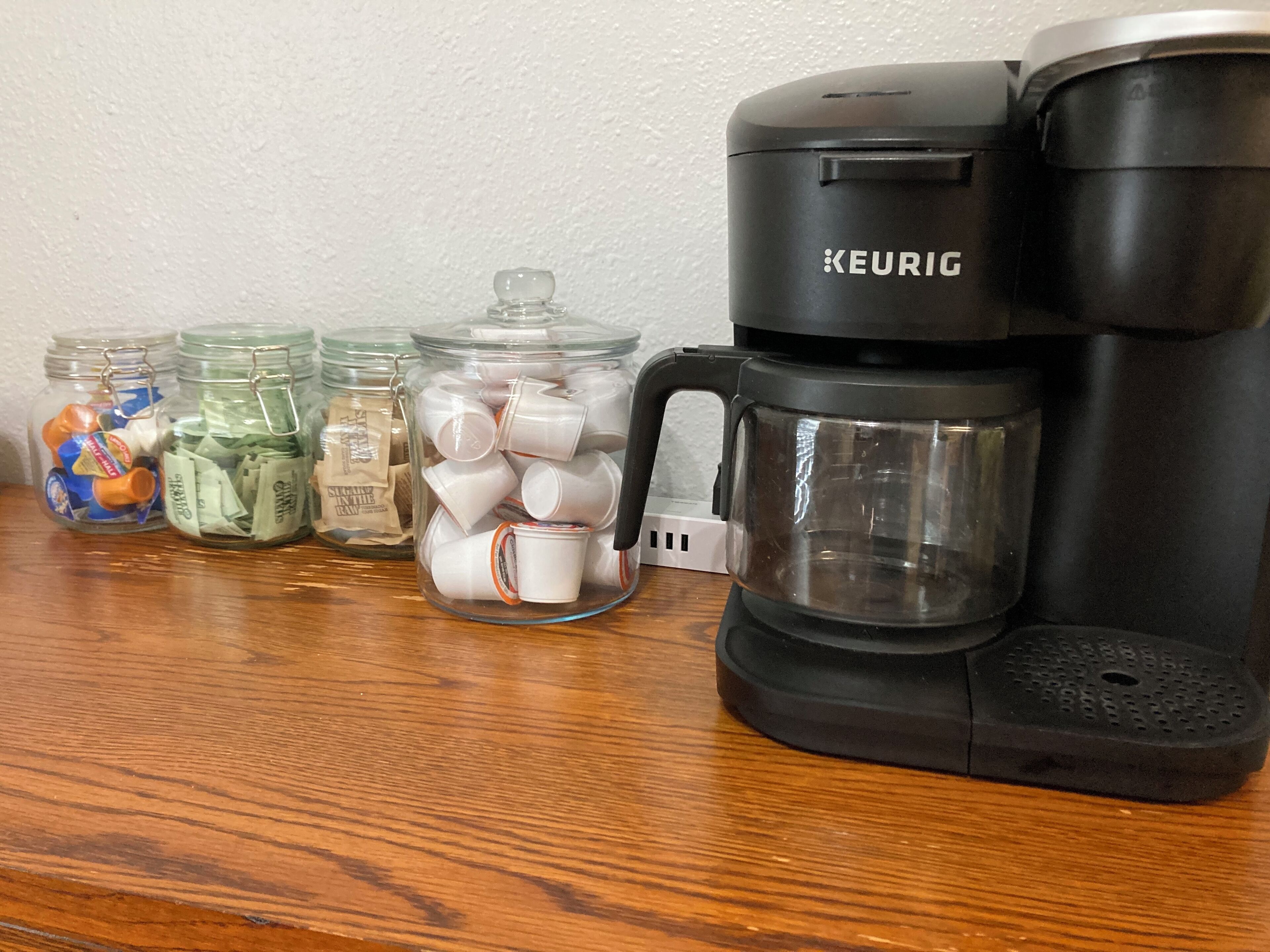 Uma cafeteira Keurig sobre uma mesa de madeira com três potes de vidro contendo diversos itens.