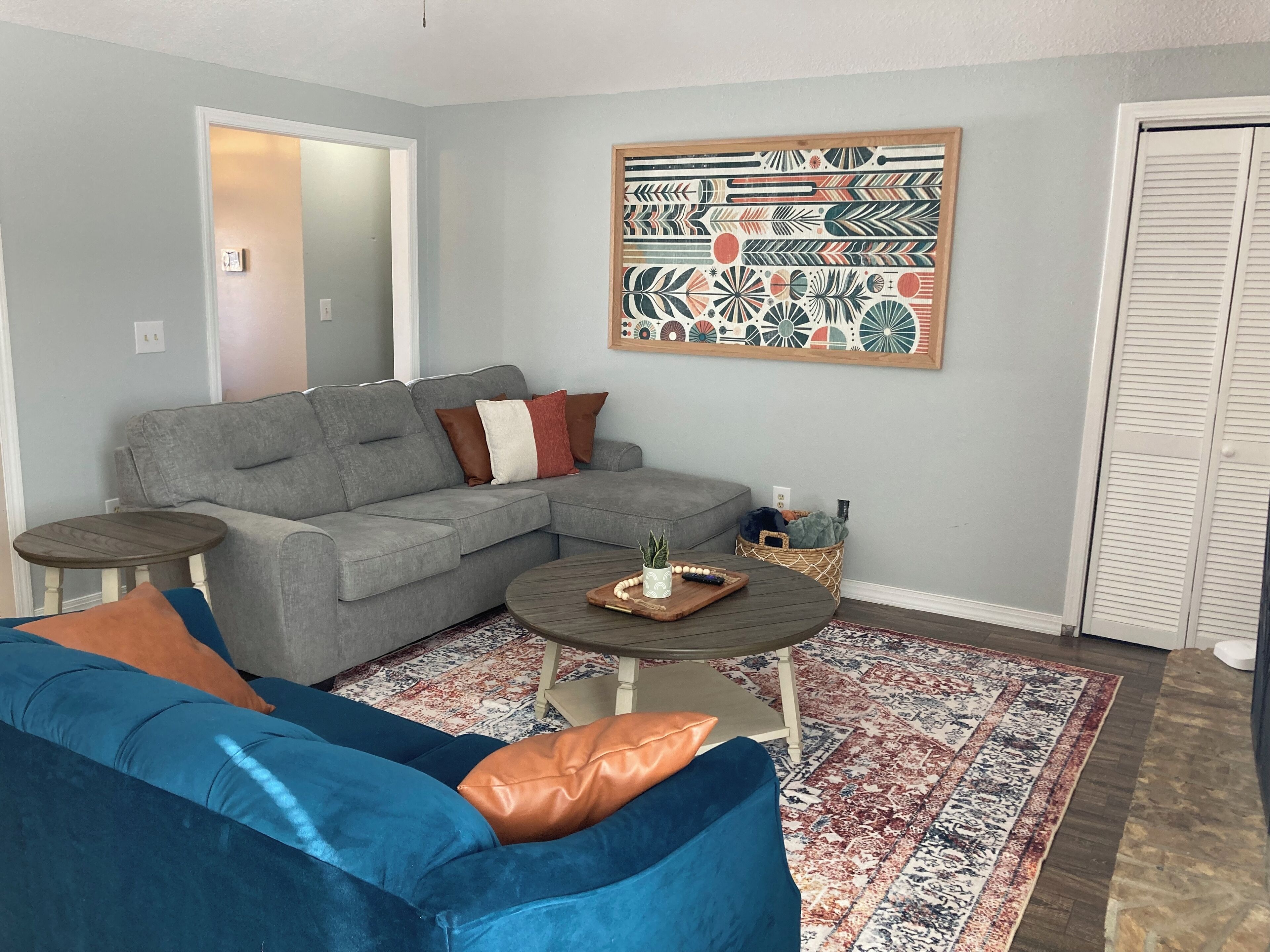 Een woonkamer met een grijze modulaire bank, een blauwe bank, een ronde salontafel en een vloerkleed met een patroon.