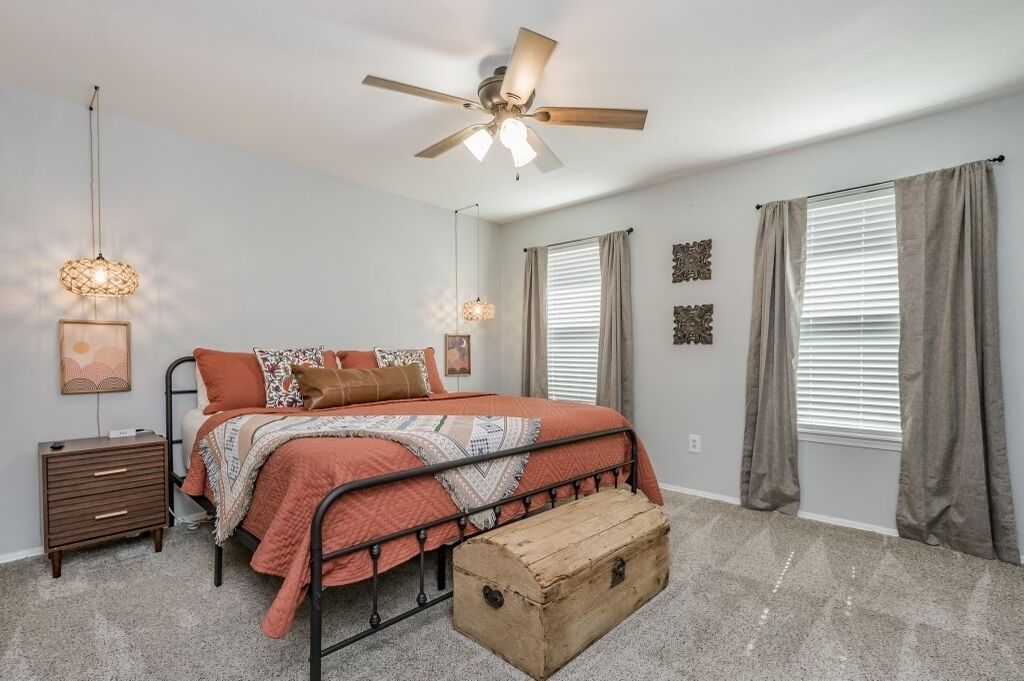 A bedroom with a bed, a nightstand, a chest of drawers, a ceiling fan, and two windows with curtains.