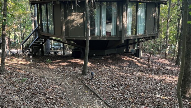 Una cabaña rústica con una escalera que sube a un porche con ventanas, rodeada por un área boscosa con suelo de tierra.