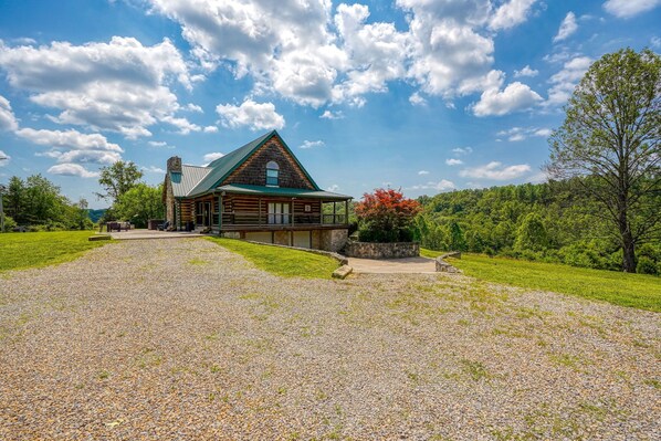 A log cabin with a green roof, surrounded by a gravel driveway and lush greenery.