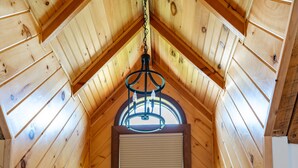 A cozy attic room with wooden walls, a window with a view of trees, a wicker chair, and a hanging light fixture. - Hidden Falls Lodge | Hocking Hills, 93 Acres (New Straitsville)