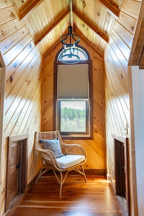 Ein gemütliches Dachzimmer mit Holzwänden, einem Fenster mit Blick auf Bäume, einem Korbsessel und einer Pendelleuchte.