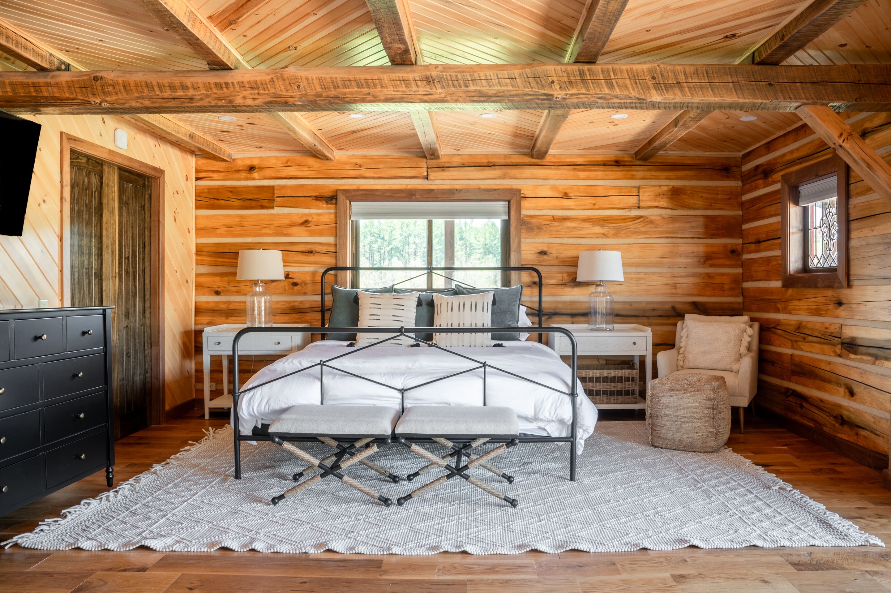 A log cabin bedroom with a metal bed frame, two bedside tables with lamps, a wooden dresser, and a window with a view of trees.