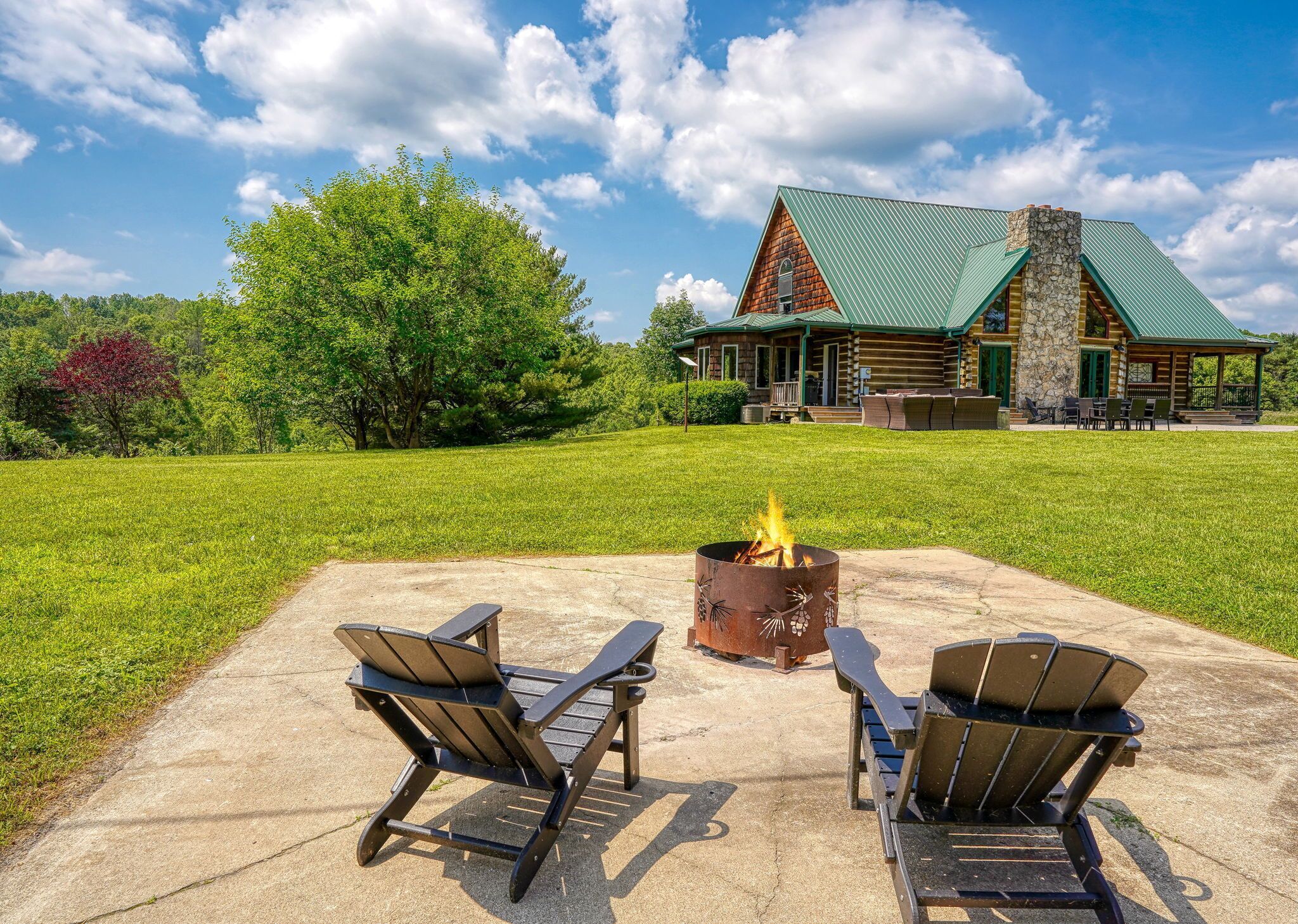 A lodge with a stone chimney, a fire pit, and two Adirondack chairs on a concrete patio.