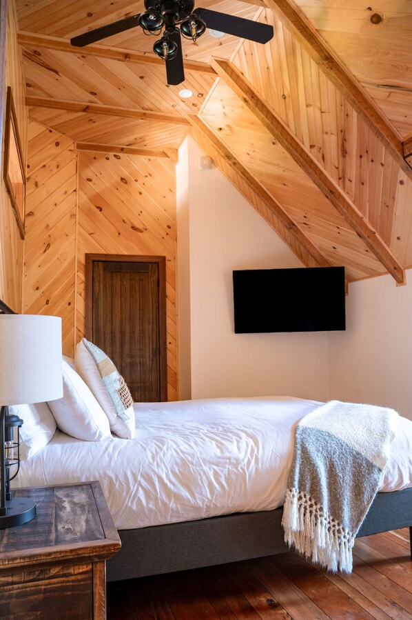 A bedroom with a wooden ceiling, a bed with white bedding, a bedside lamp, and a TV mounted on the wall.