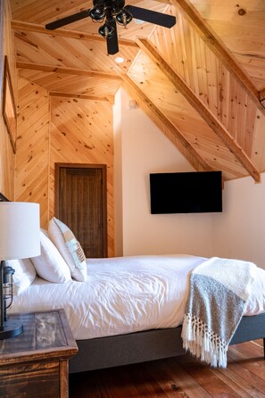 Un dormitorio con techo de madera, una cama con ropa de cama blanca, una lámpara de noche y un televisor montado en la pared.