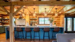 A rustic kitchen with wooden beams, a marble island with bar stools, hanging pendant lights, and a large window. - Hidden Falls Lodge | Hocking Hills, 93 Acres (New Straitsville)