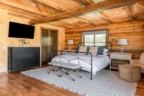 Ein Schlafzimmer in einer Blockhütte mit einem Metallbettgestell, einem Schminktisch, einem Nachttisch und einem Fenster mit Blick auf Bäume.
