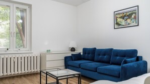 A modern living room with a blue sofa, a glass coffee table, a white radiator, and a window with blinds. - Koszutka Blue Apartment (Katowice)