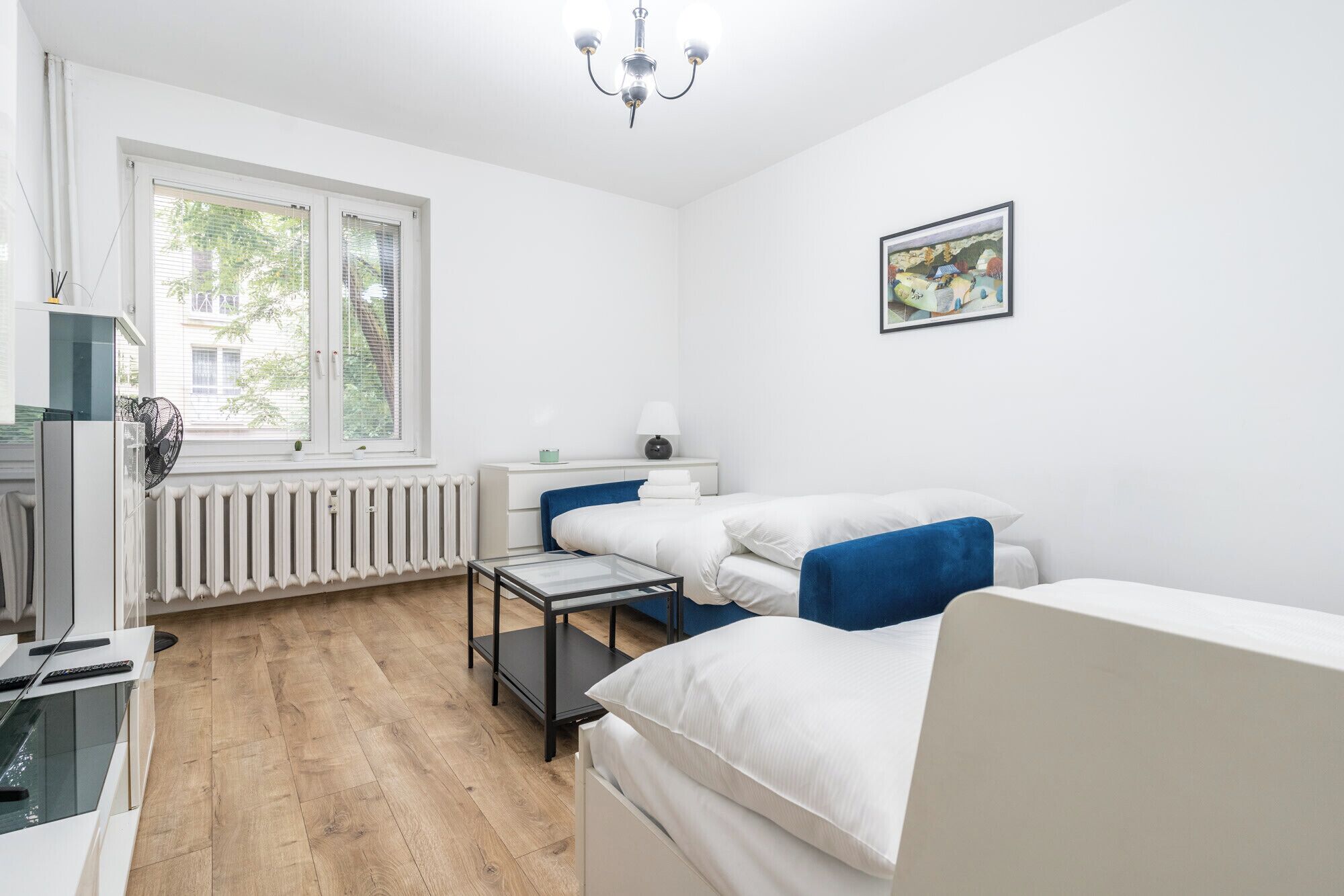 A modern, well-lit bedroom with a large window, a radiator, a desk, and a bed with blue pillows.