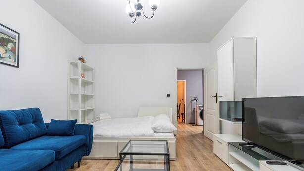 A modern living room with a blue sofa, a glass coffee table, a bed with white bedding, a TV on a white cabinet, and a white shelving unit. - Koszutka Blue Apartment (Katowice)