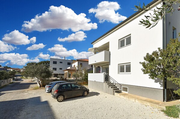 A residential building with a white exterior, multiple windows, and a visible balcony. There is a car parked in front and a gravel driveway.